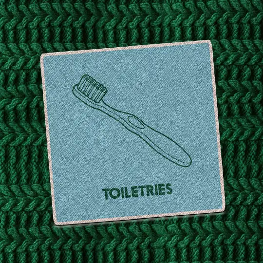 Teal fabric patch with a dark green illustration of a toothbrush and the words “Toiletries” in bold capital letters, placed on a chunky dark green knit background.